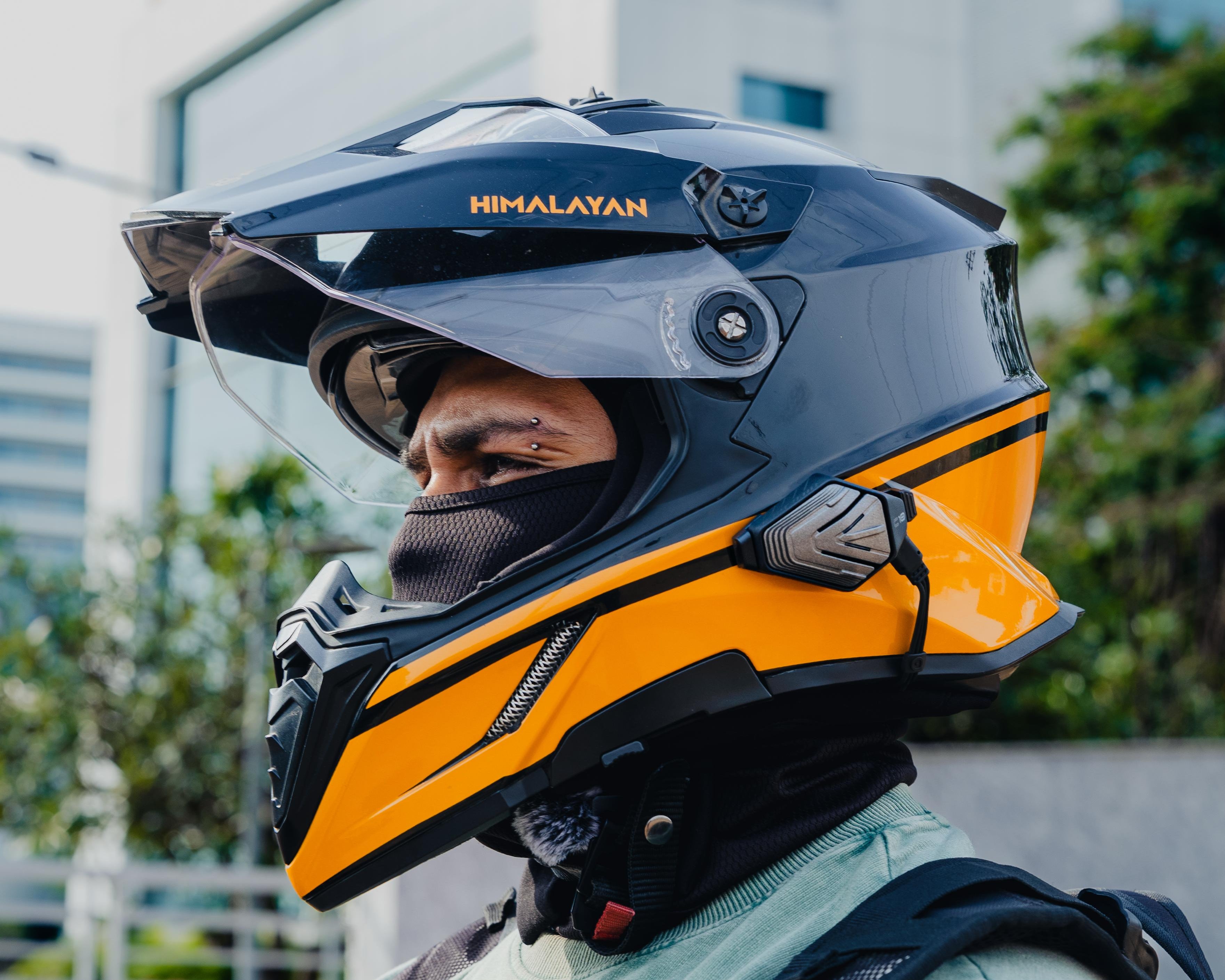 A person wearing a full-face motorcycle helmet in black and bright orange, with the brand name HIMALATAN visible on the side, photographed outdoors in front of a modern building.